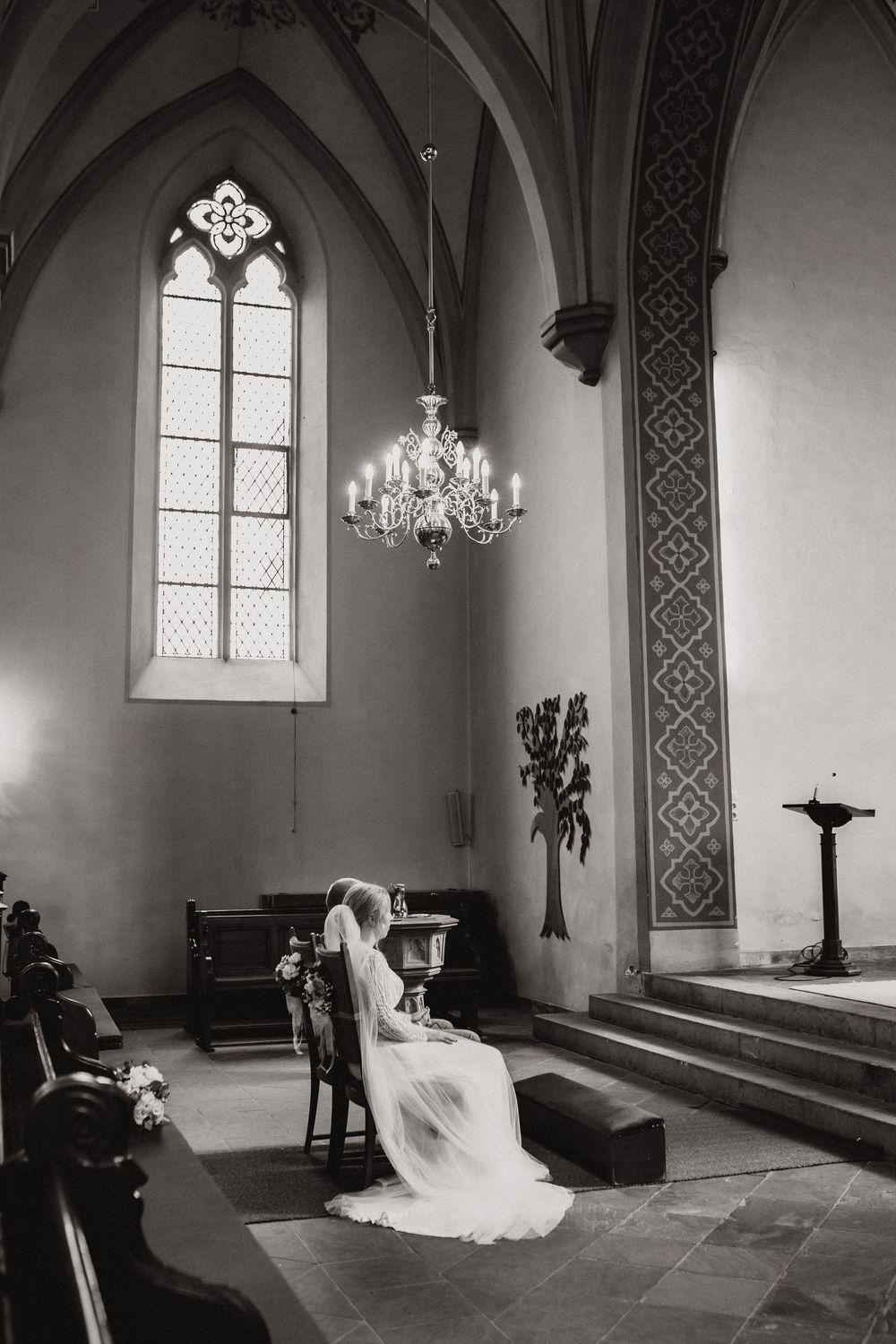 Braut im weißen Hochzeitskleid mit langem Schleier sitzt in stiller Andacht in einer gotischen Kirche mit Lüster – Hochzeitsfotografie Julia Kippes